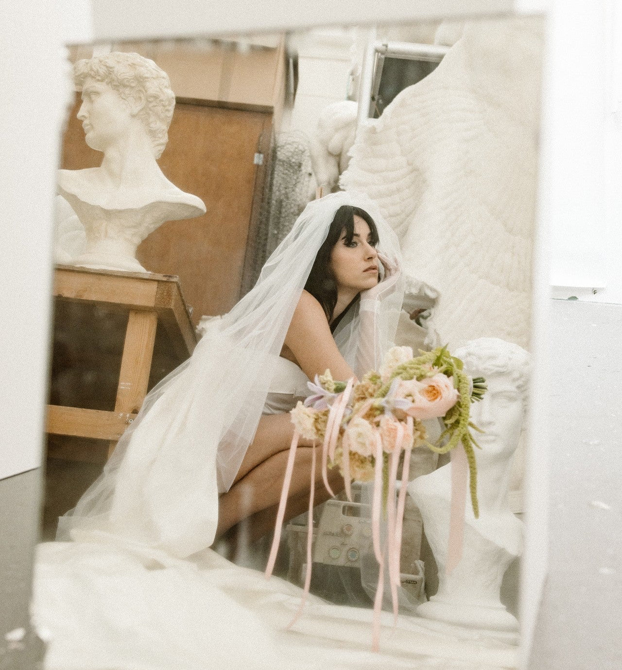 Woman in a wedding dress sitting among marble sculptures with flowers.
